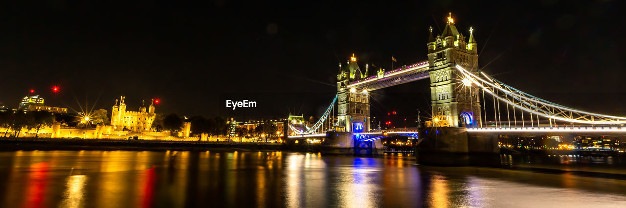 ILLUMINATED BRIDGE OVER RIVER WITH CITY IN BACKGROUND AT NIGHT