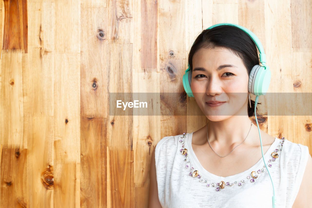 Portrait of smiling woman listening to music through headphones against wooden wall