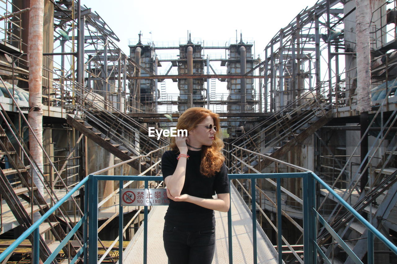 PORTRAIT OF YOUNG WOMAN STANDING ON RAILING AGAINST BUILDINGS