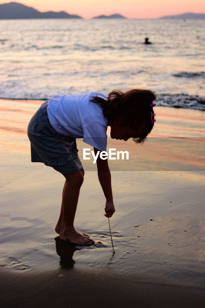 Side view full length of girl bending on shore at beach during sunset