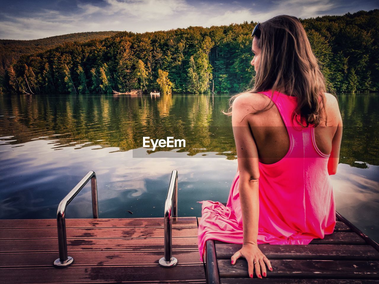 Rear view of woman sitting on lounge chair by lake against sky
