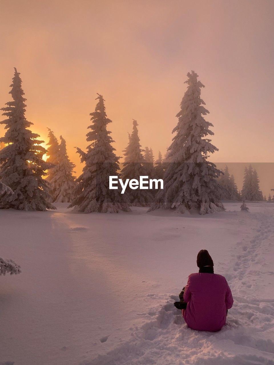 rear view of woman sitting on snow covered landscape during sunset