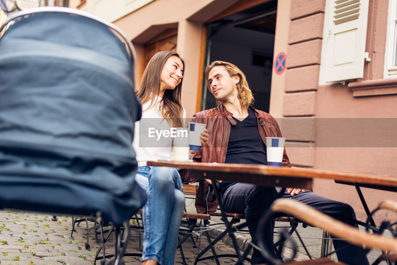 Smiling couple with baby stroller drinking coffee at sidewalk cafe 