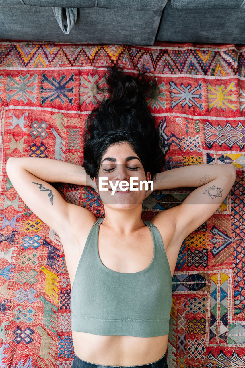 Top view of cheerful hispanic woman in sports bra holding hand behind head and smiling with closed eyes while lying on ornamental carpet near sofa during break in training at home