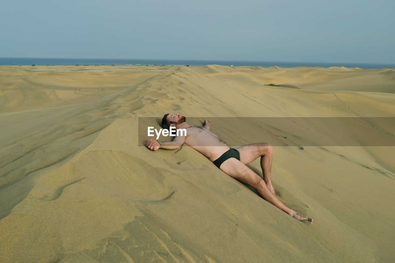 Full length of shirtless man lying down on sand at beach against sky