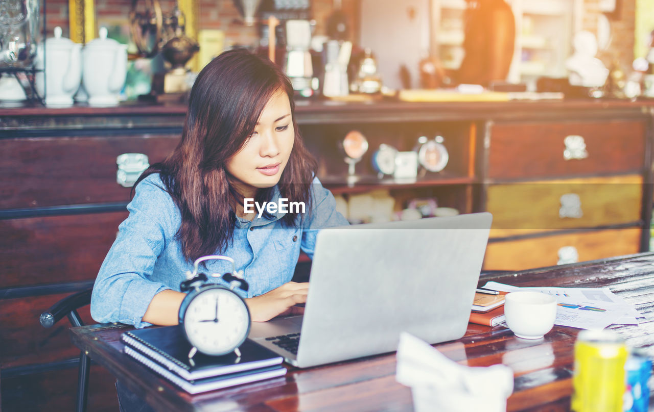 YOUNG WOMAN USING PHONE WHILE SITTING IN LAPTOP