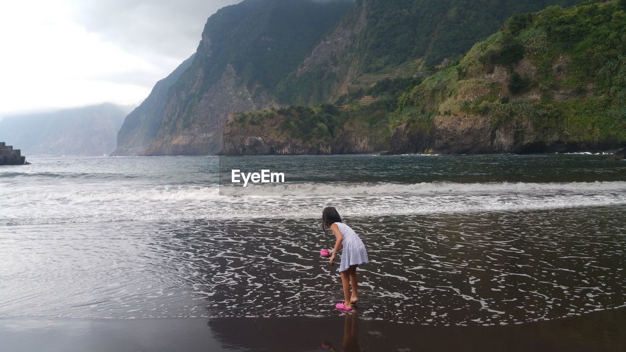 Rear view of girl on shore at beach against rocky mountains