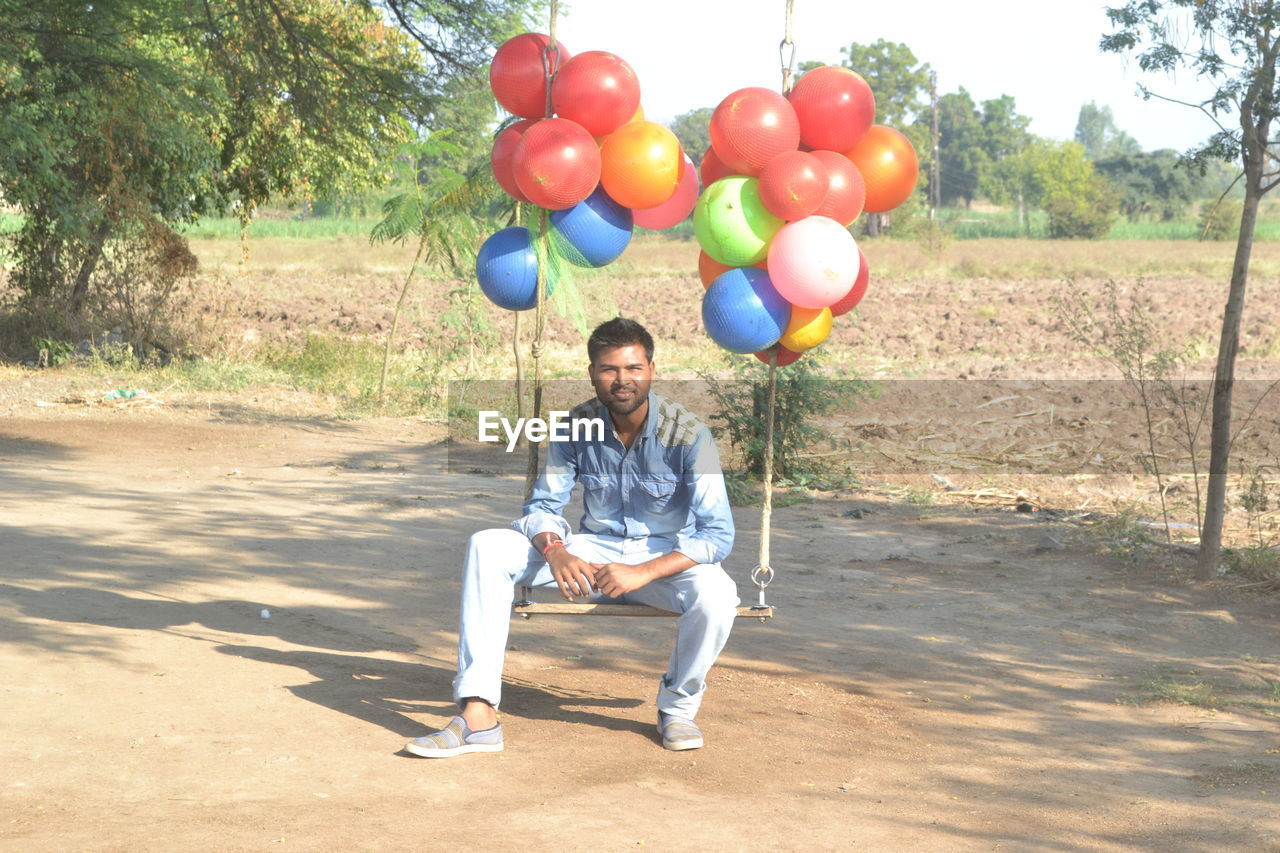 Man sitting on swing with colorful balloons against field