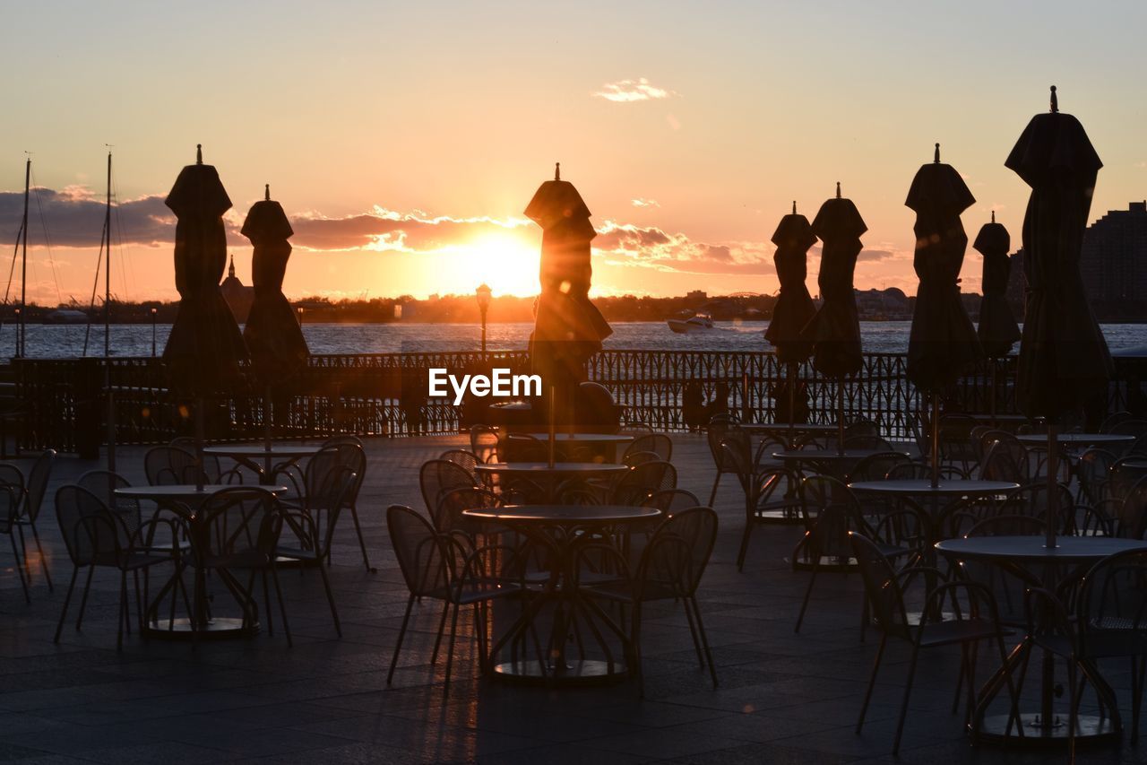 Empty chairs and tables by closed parasols at outdoor cafe during sunset