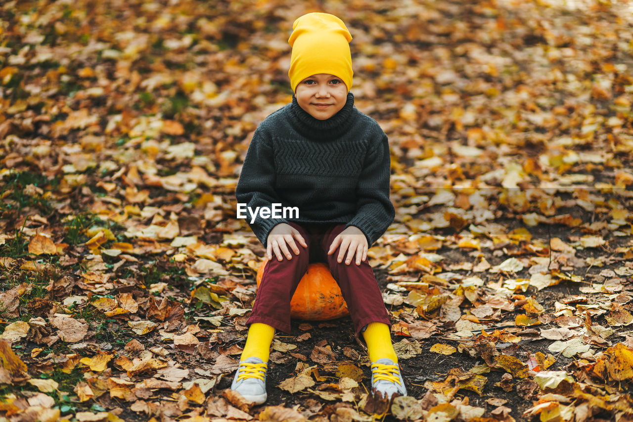 A boy a teen child in a bright yellow hat sits on a pumpkin in an autumn forest in nature outdoors