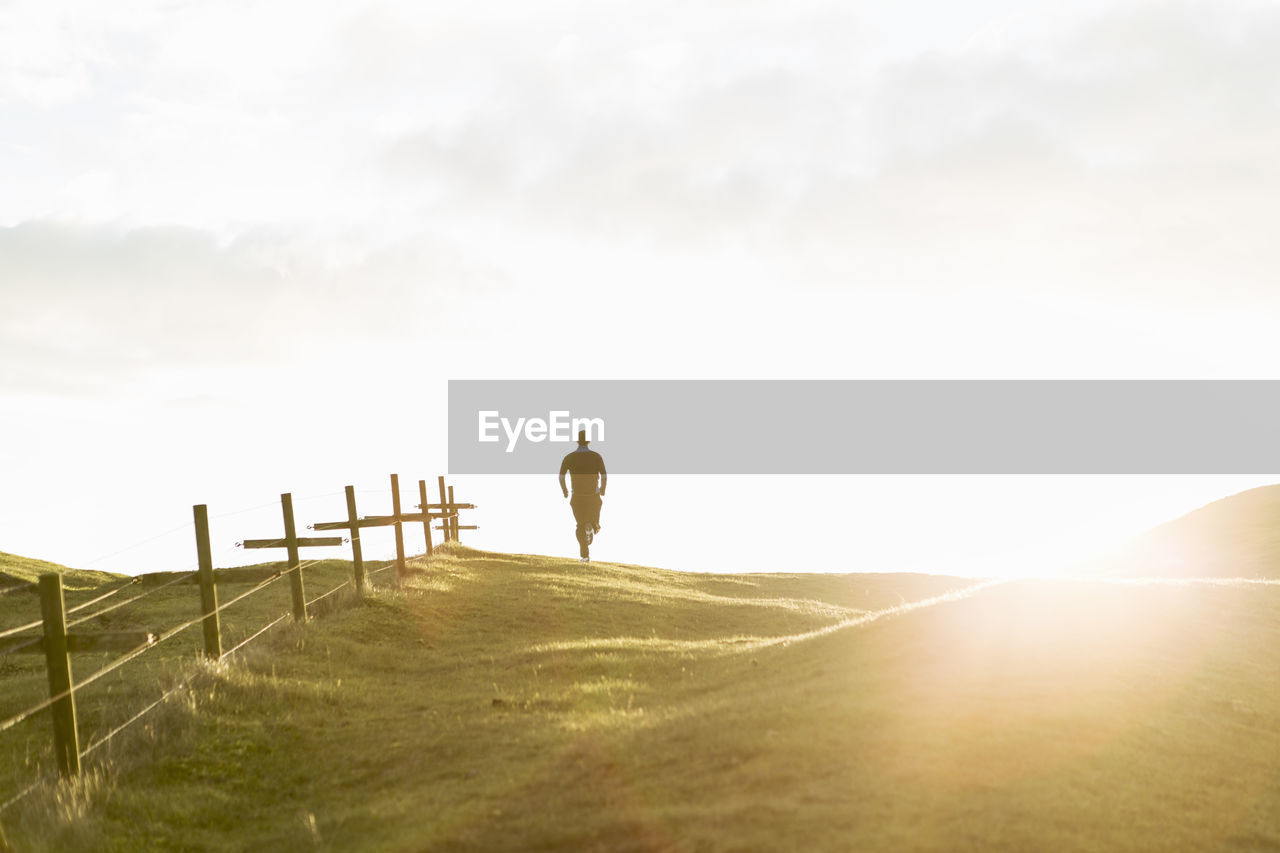 Distant view of man jogging on grassy field against sky during sunny day