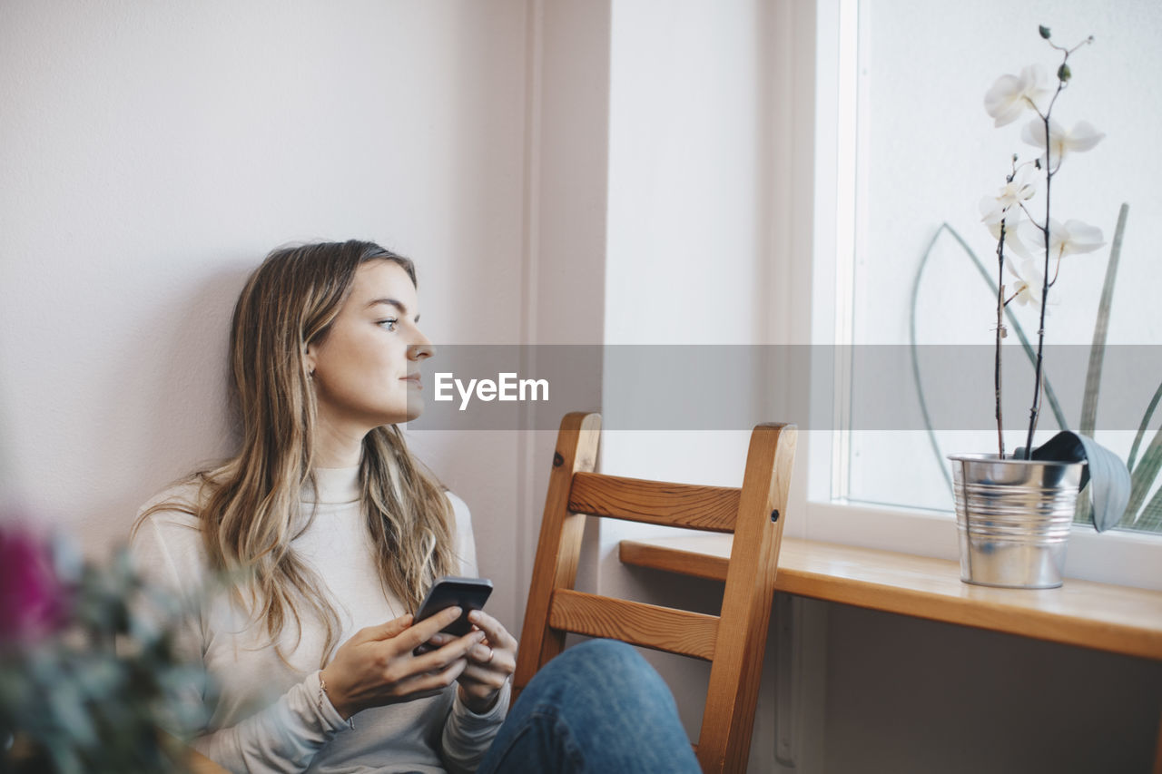 Young woman sitting with mobile phone on chair while looking through window at dorm