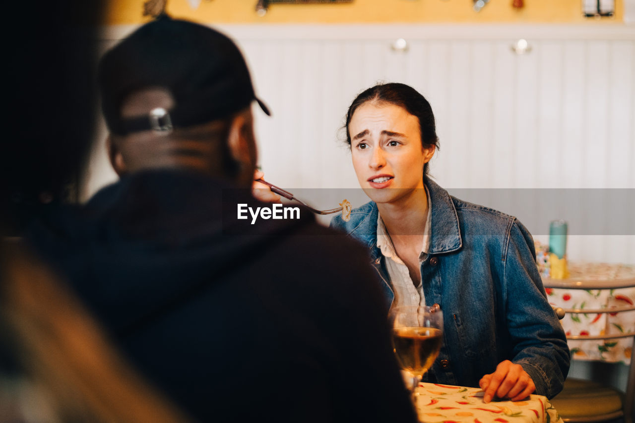 Young woman eating while talking with male friend sitting at restaurant during brunch