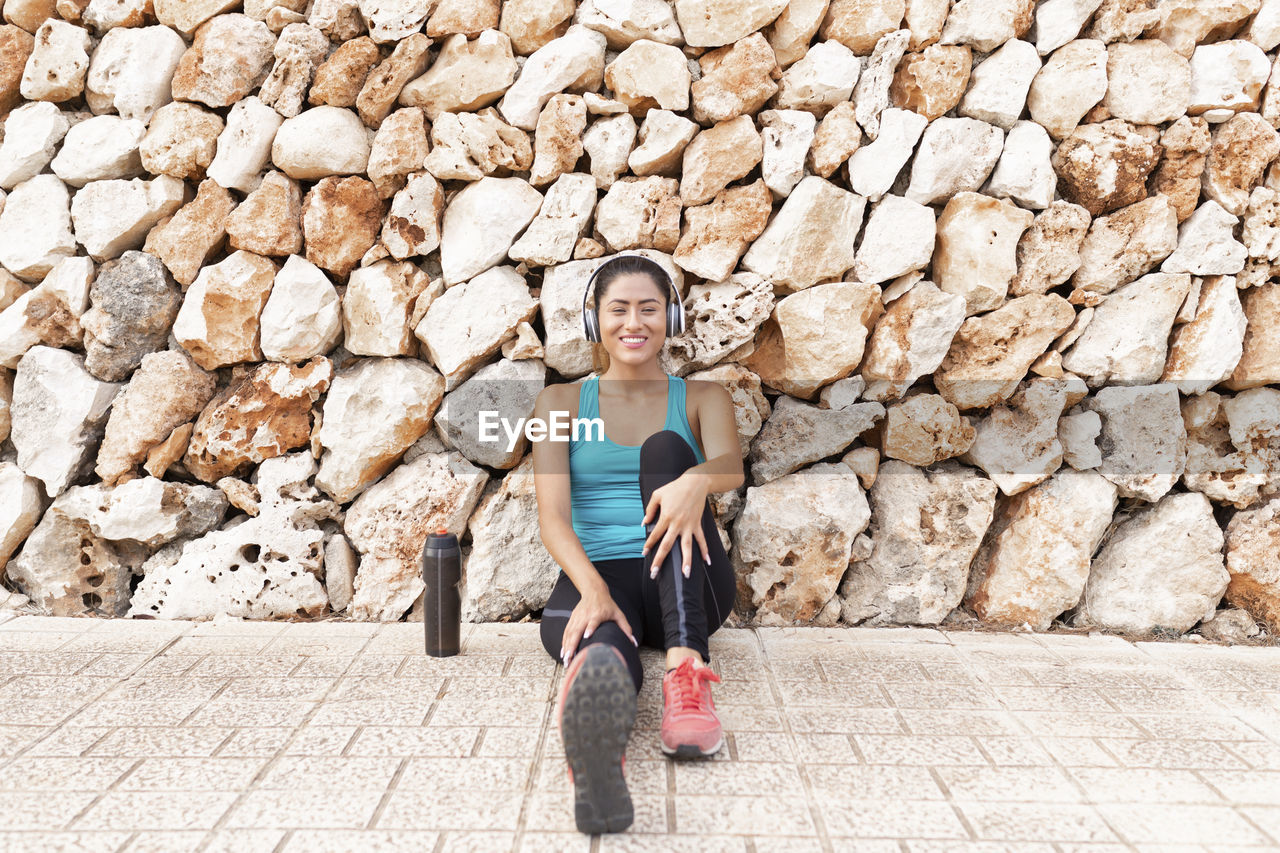 Smiling beautiful young woman listening music through headphones while sitting against stone wall after exercising