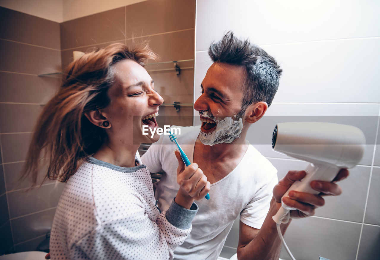 Cheerful man holding dryer for woman in bathroom