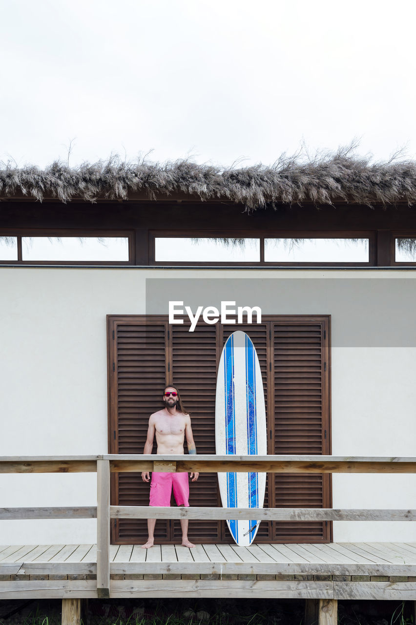 Young man standing by surfboard in front of door