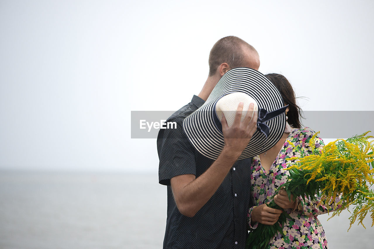 Young romantic couple kisses and hugging, hiding their faces behind their summer hats