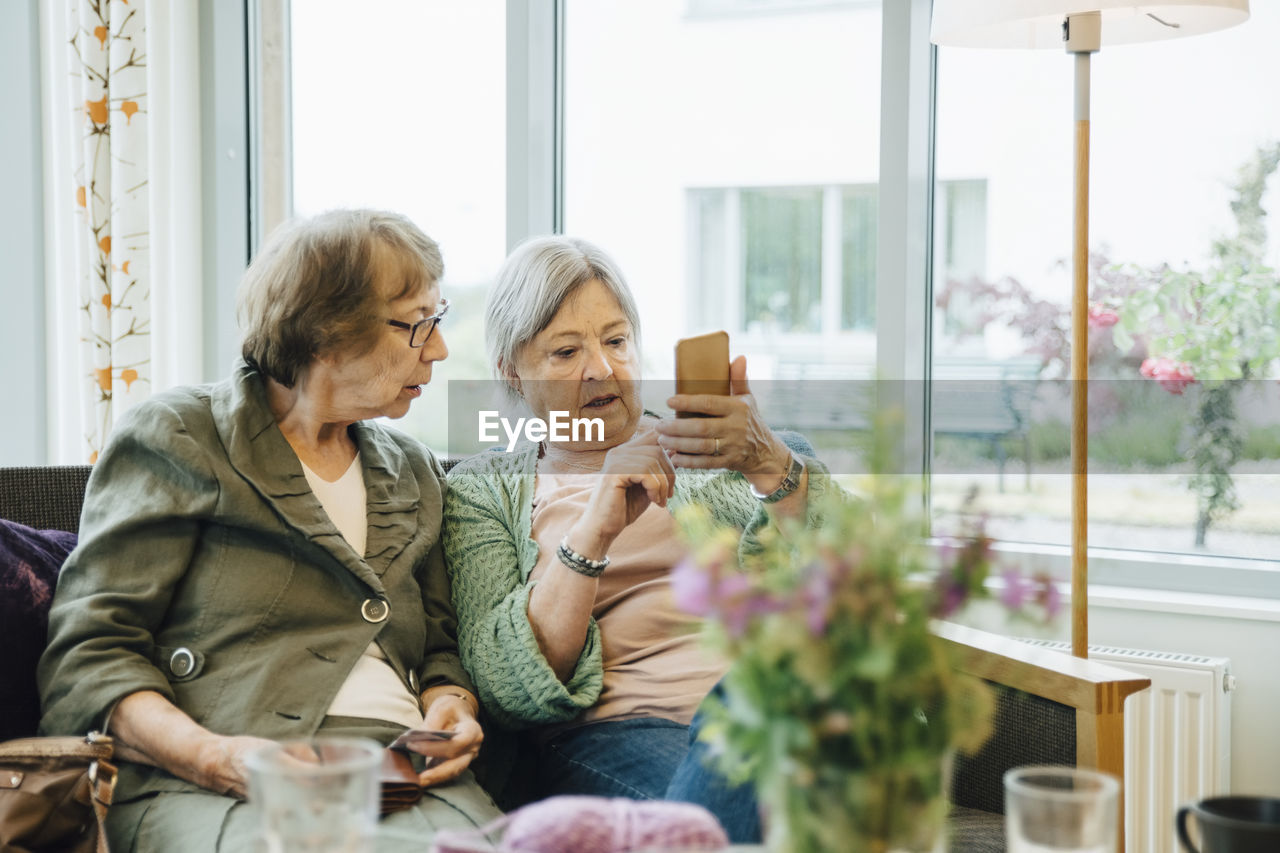Elderly woman using mobile phone while sitting with female friend on sofa against window at retirement home