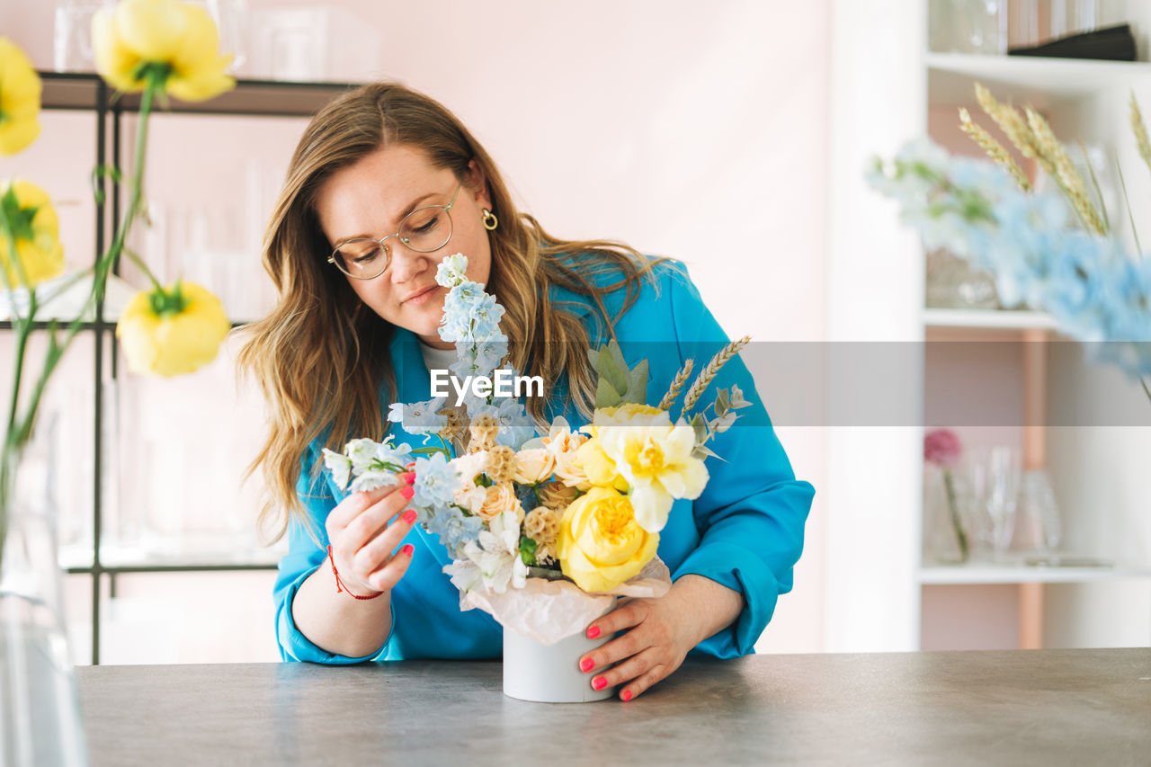 Young woman florist in blue suit and eye glasses with bouquet of flowers in box in flower shop