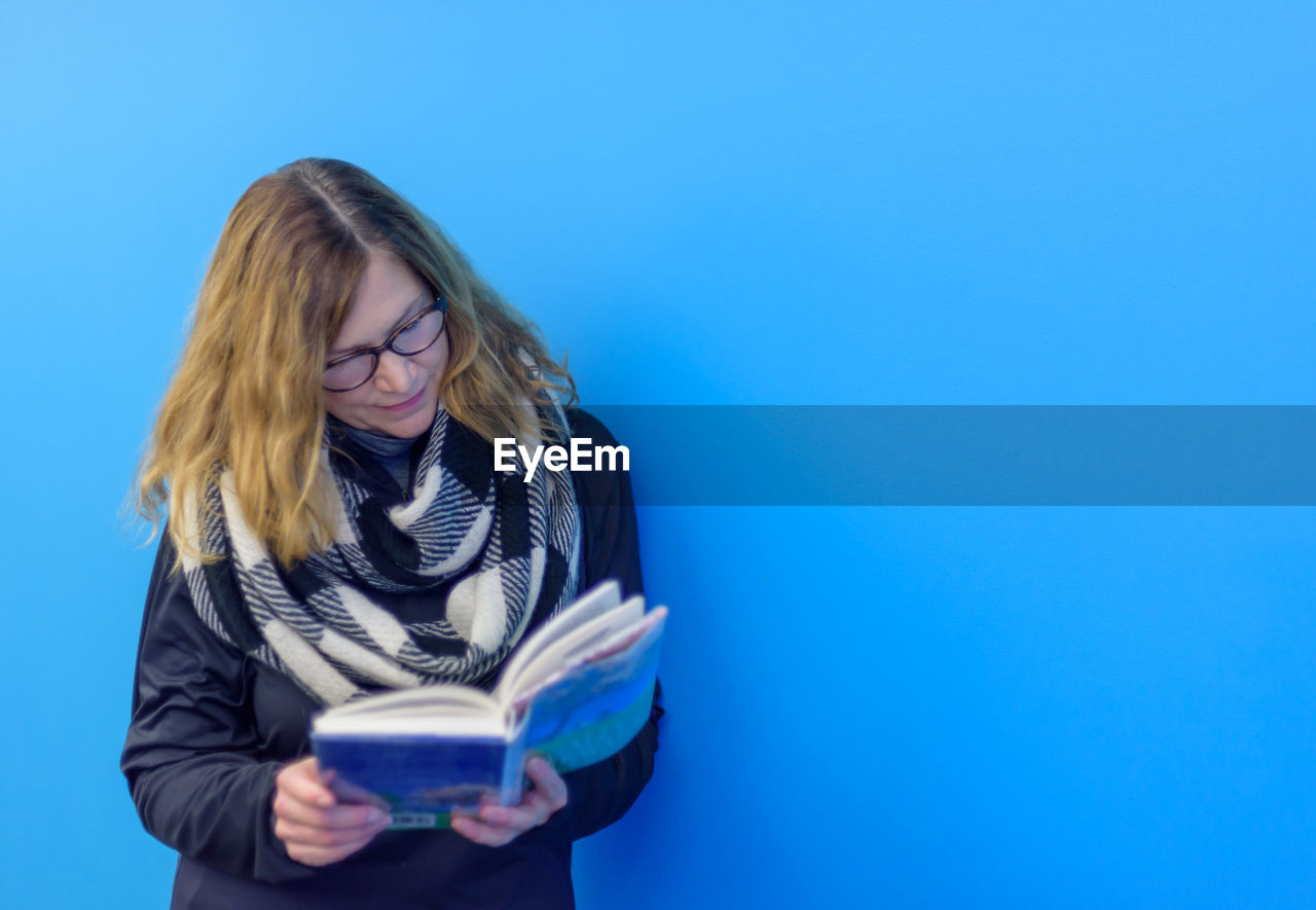 Mature woman reading book while standing against blue background