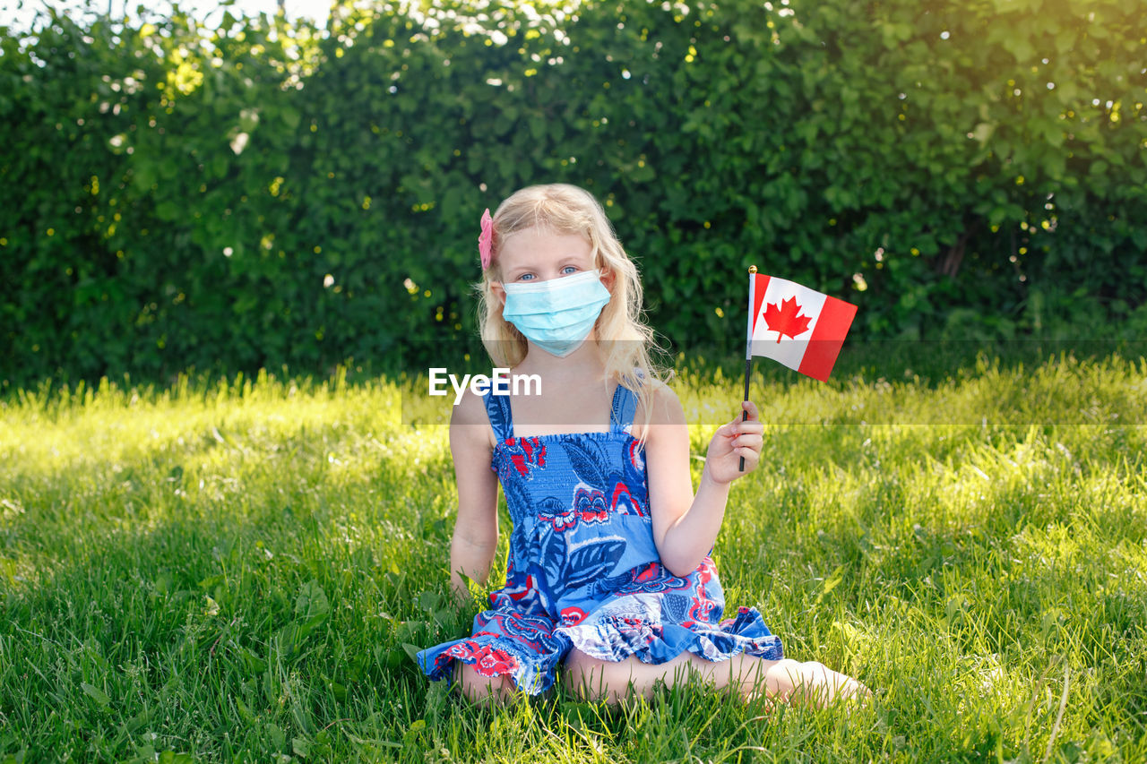 Portrait of girl wearing mask holding canadian flag sitting on grass