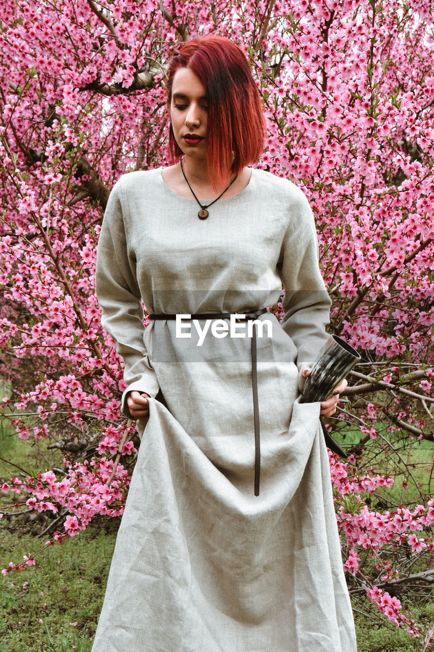 spring, plant, women, one person, flower, adult, flowering plant, pink, young adult, nature, springtime, beauty in nature, blossom, happiness, smiling, redhead, tree, freshness, standing, female, fashion, lifestyles, clothing, portrait, dress, growth, emotion, hairstyle, front view, gown, outdoors, three quarter length, looking, cheerful, fragility, day, cherry blossom, leisure activity, formal garden, garden