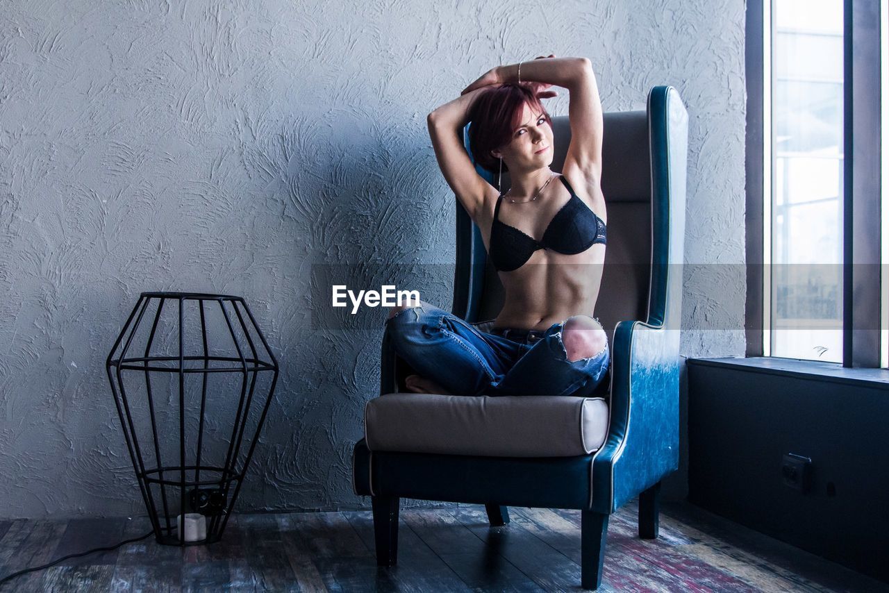 Portrait of woman wearing bra while sitting on armchair by window at home