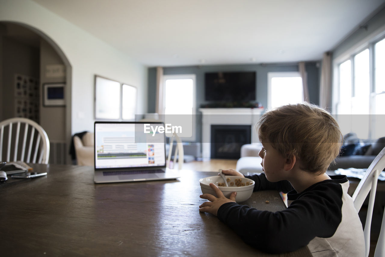 Wide side view of blonde boy eating cereal watching lesson on laptop