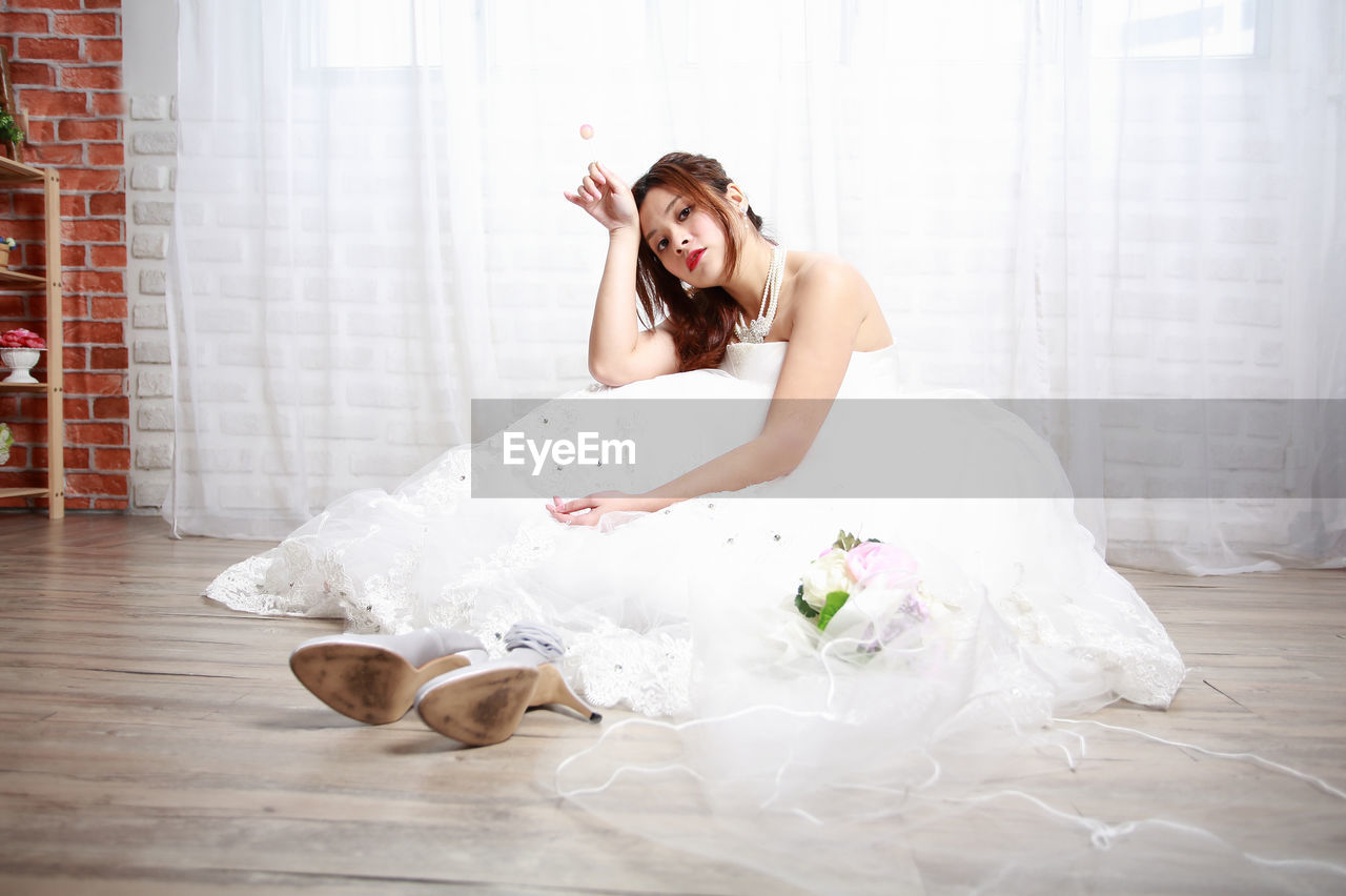 Portrait of bride eating lollipop while sitting on hardwood floor against curtain