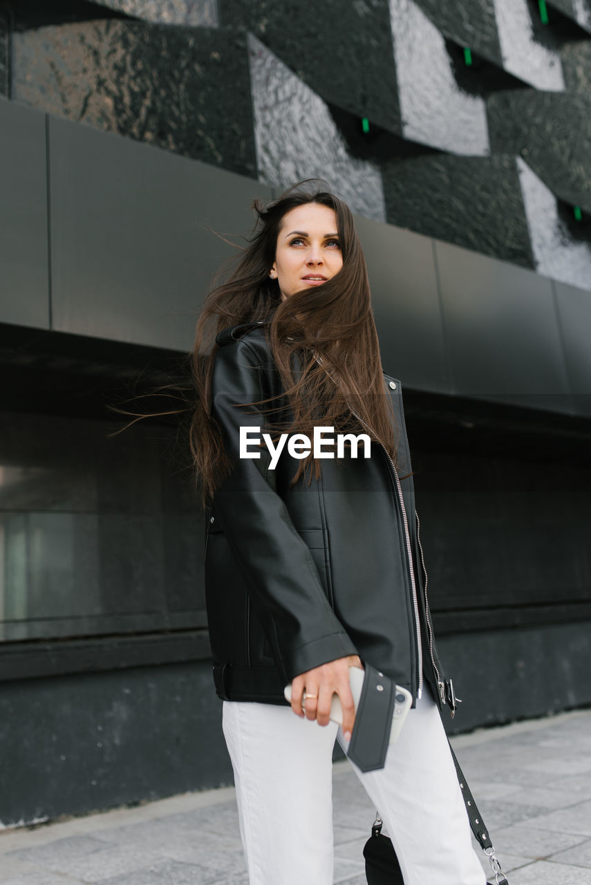 Stylish woman with long hair is standing in front of building, wearing black jacket and white pants