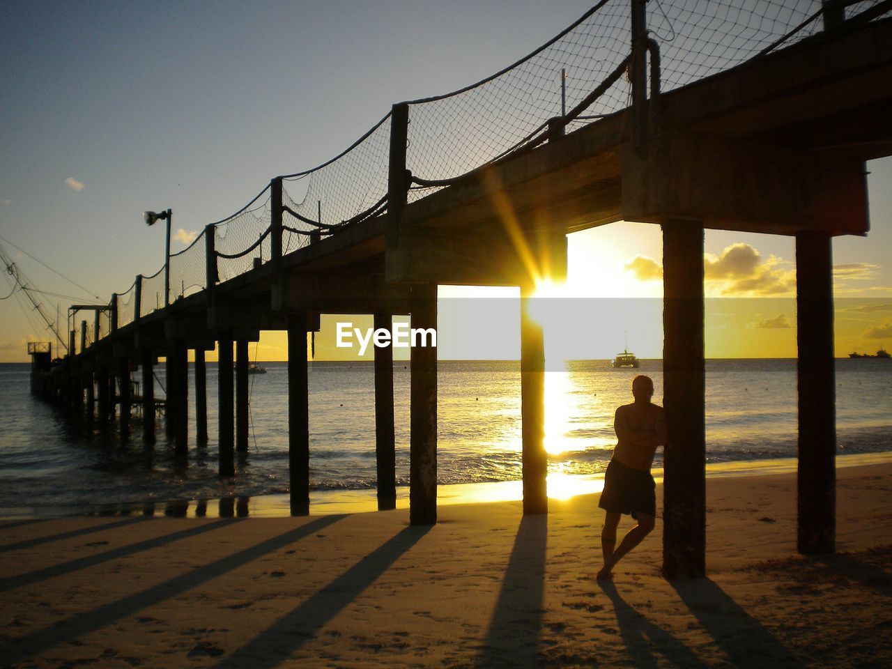 Full length of man standing by pier at beach against clear sky during sunset