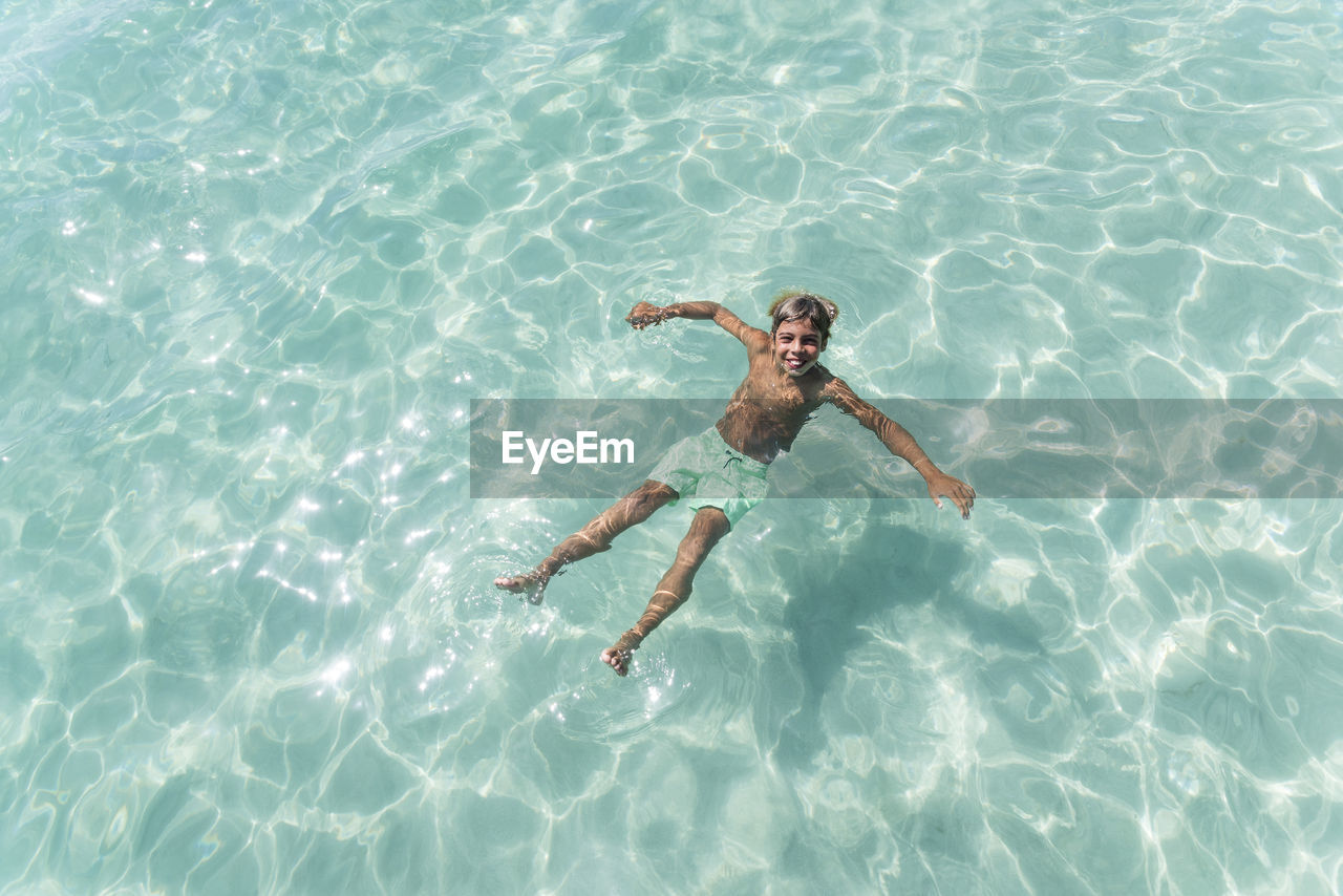 From above of delighted teenage boy swimming in clear sea water on sunny day and looking at camera while enjoying summer vacation