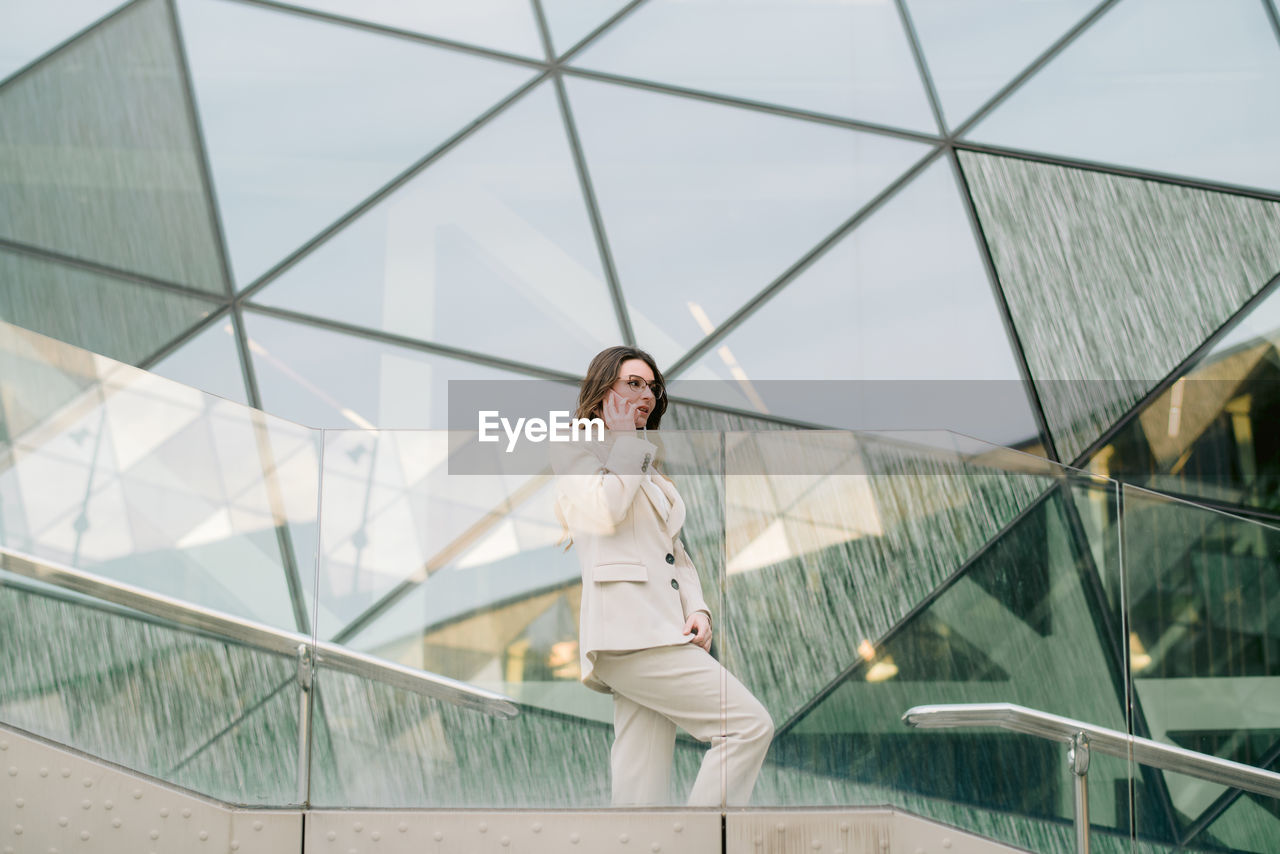 Side view low angle of determined female entrepreneur in elegant suit standing near glass building in downtown and talking on mobile phone while discussing work issues and looking away