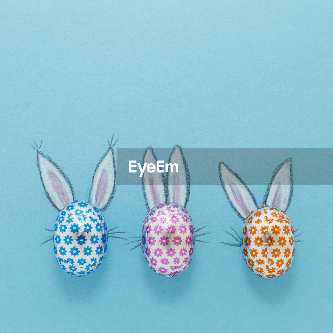 Three chocolate eggs in colorful aluminium foil with painted easter bunny ears on blue background