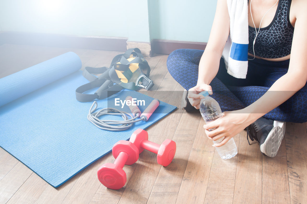 Low section of woman with water bottle by dumbbells on hardwood floor