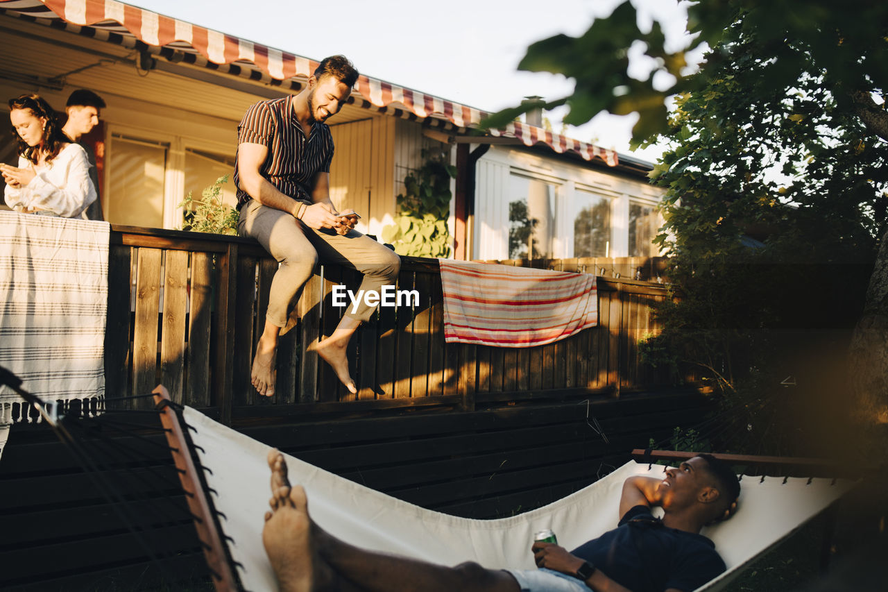 Man lying in hammock talking to friend sitting on railing in back yard