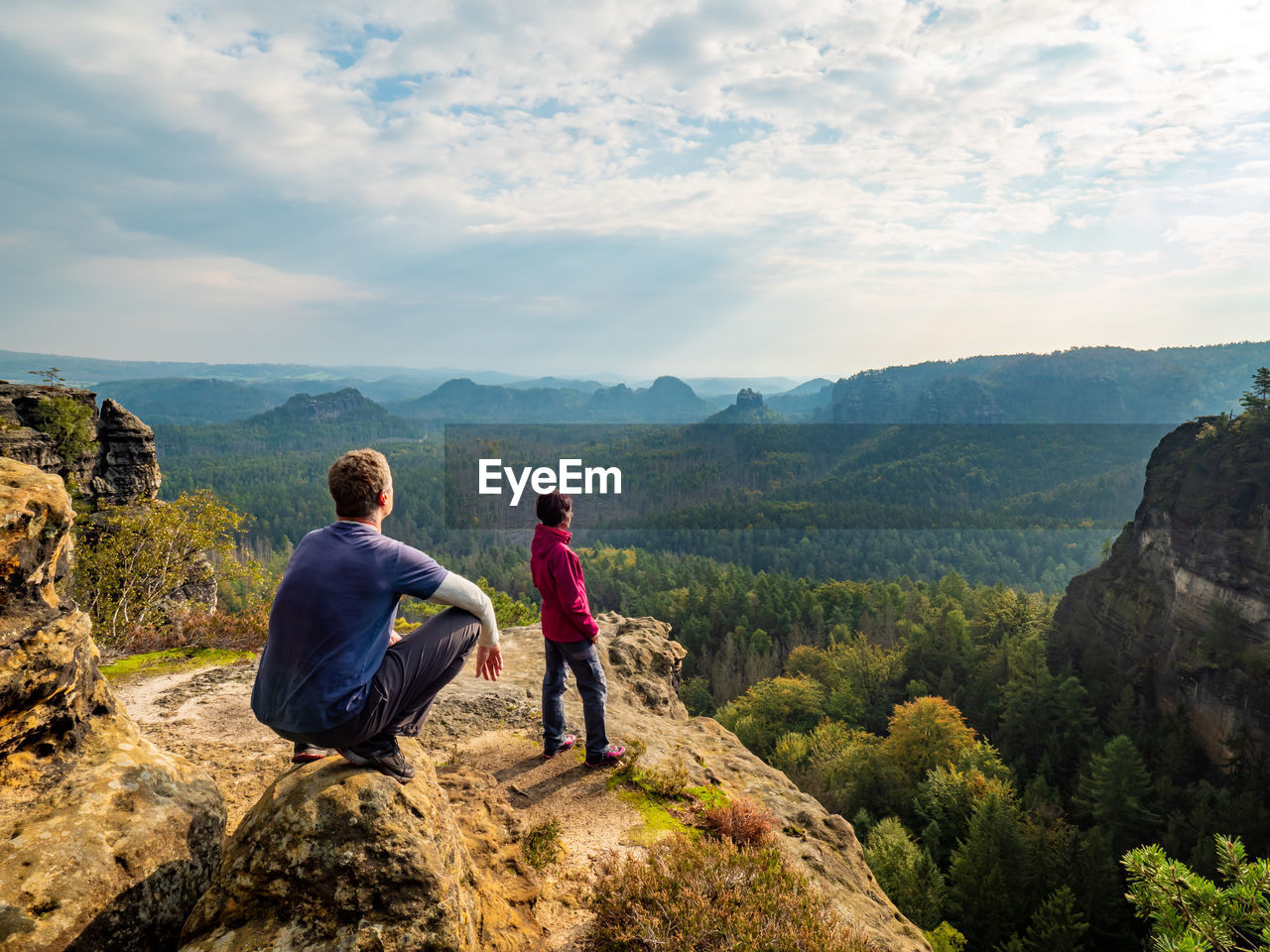 Man hiker sit on edge and woman tourist bellow watching over sunny landscape saxon switzerland.