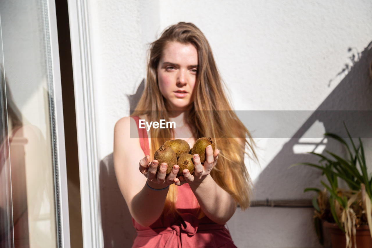 Portrait of young woman holding kiwis against wall