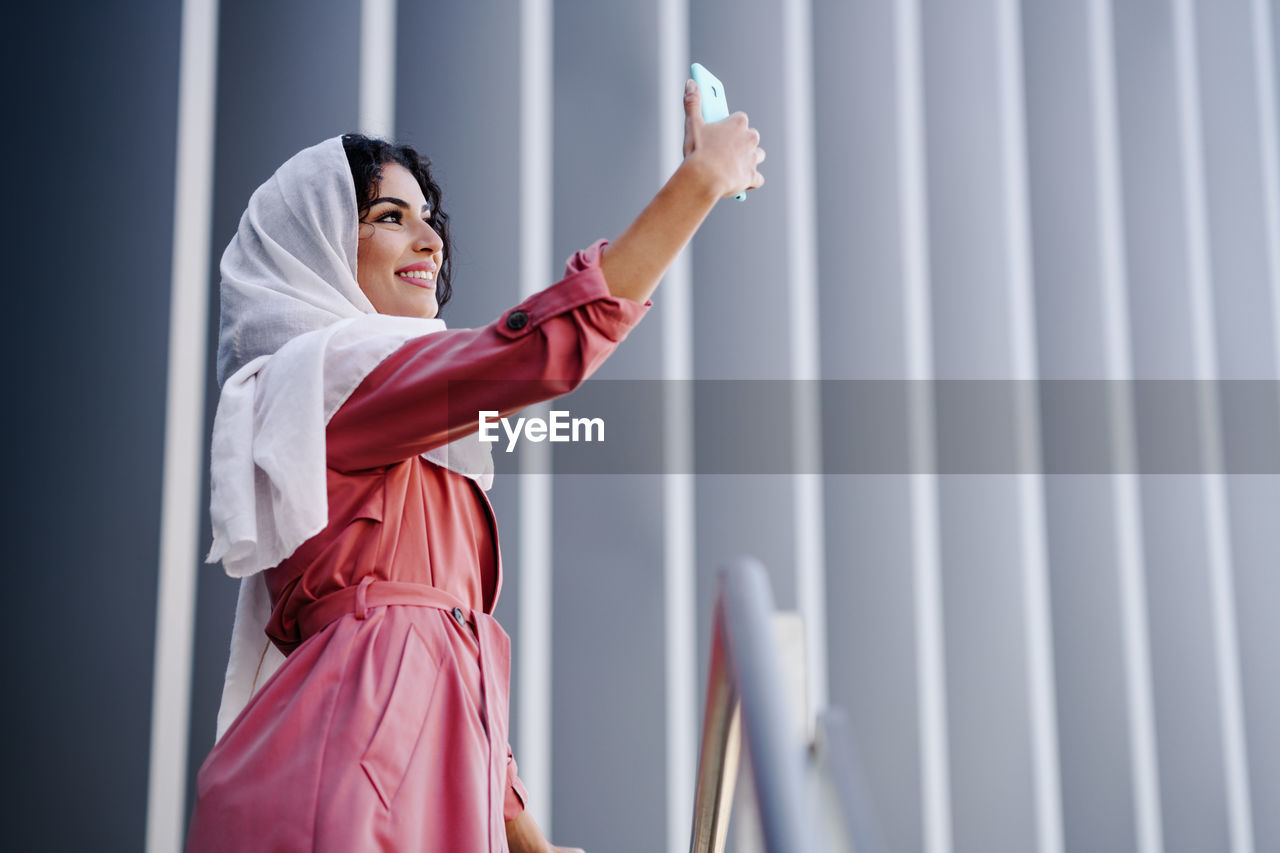 Woman taking selfie while standing against wall