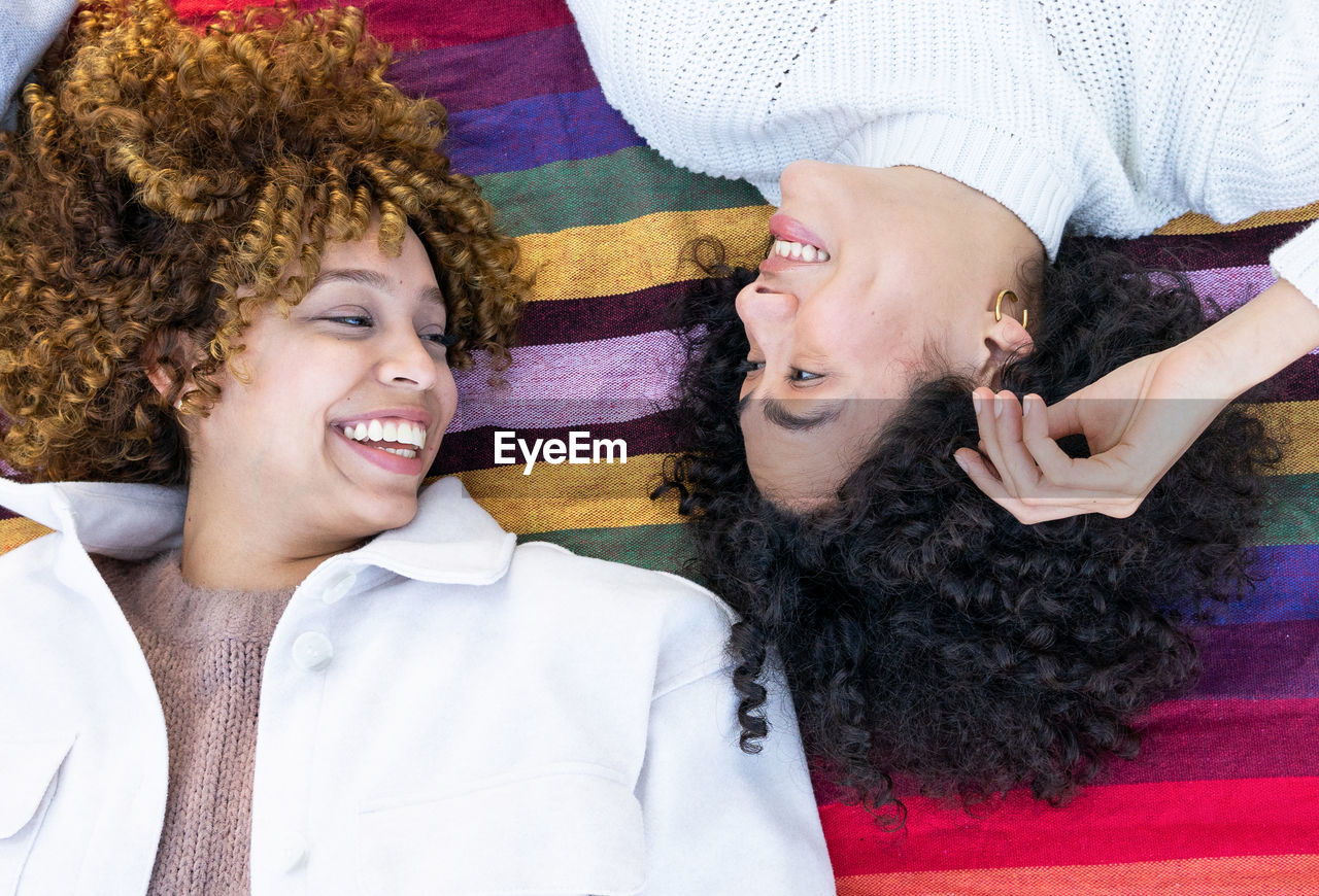 From above of happy multiracial girlfriends with curly hair lying on colorful plaid in park and looking at each other while resting together