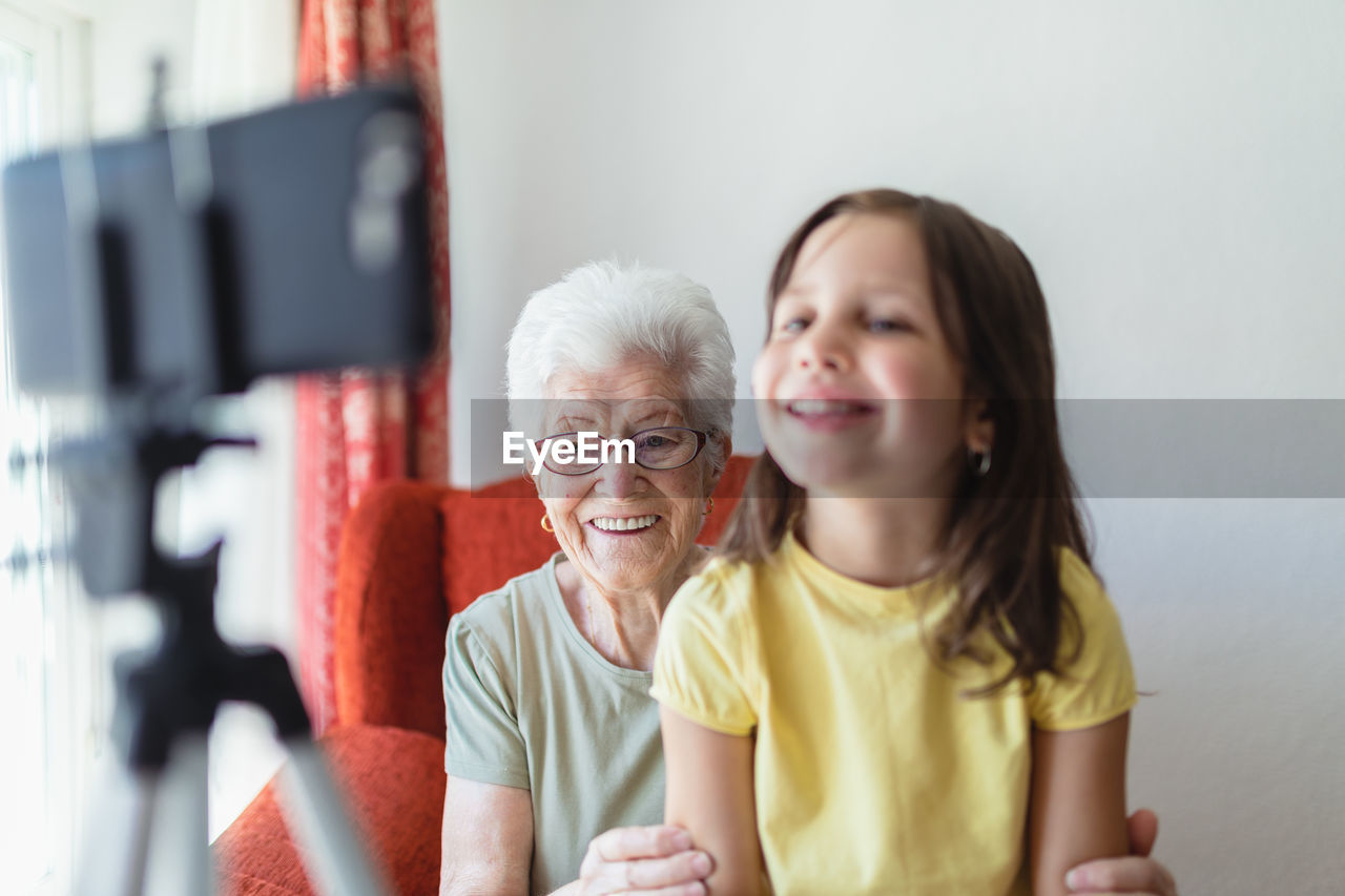 Smiling senior woman and girl sitting in living room together and having video call on smartphone