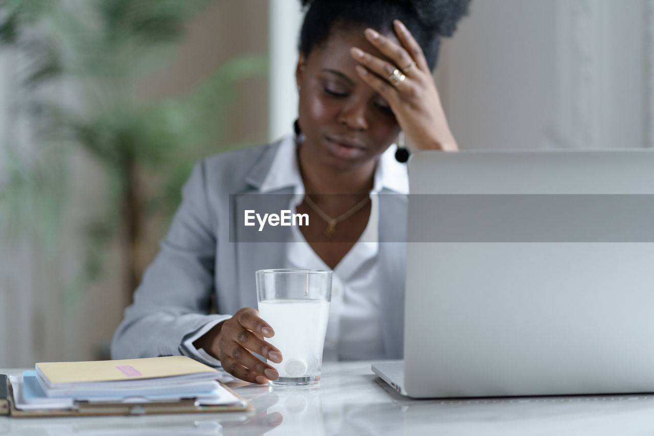 Ill woman holding glass with medicine by laptop on table