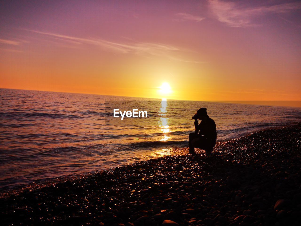 Side view of silhouette man photographing on shore at beach against dramatic sky during sunset