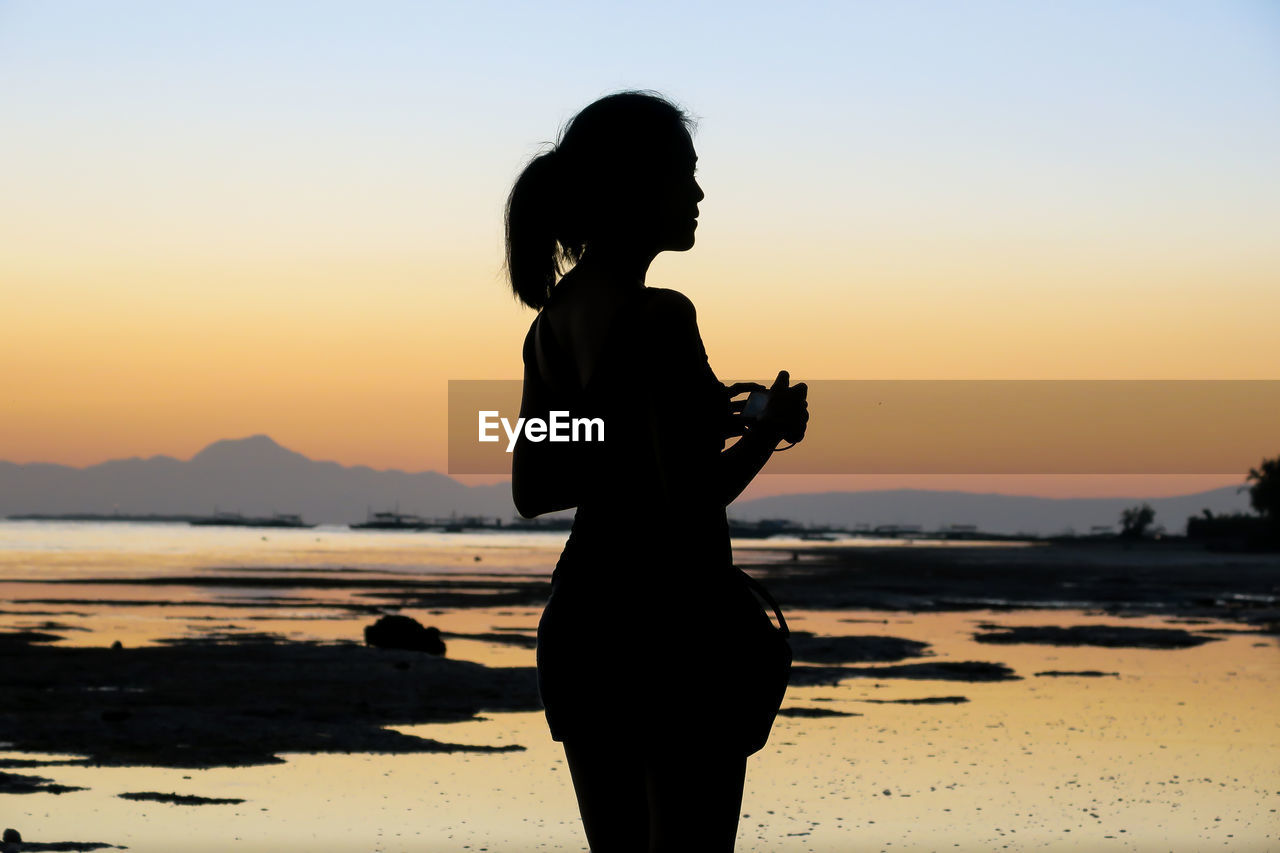 Silhouette woman standing at beach against clear sky during sunset