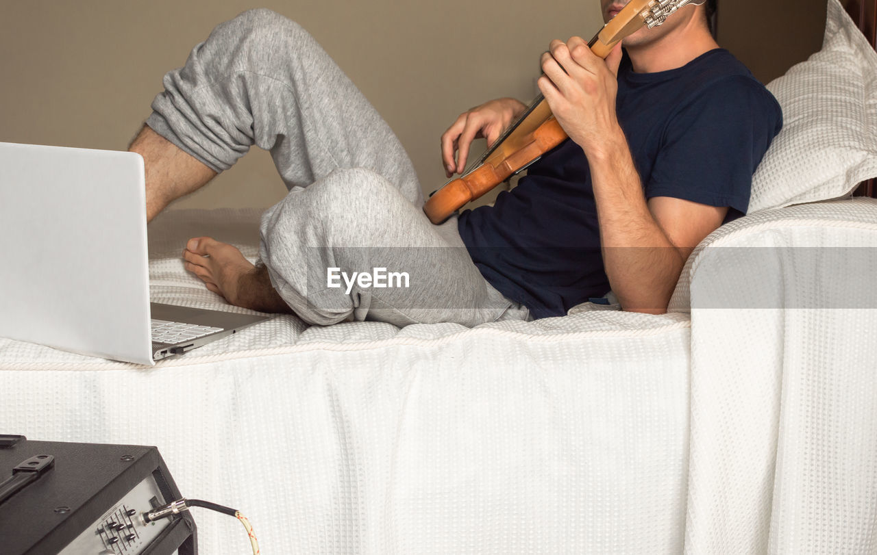 Young man lying on a bed playing electric bass.
