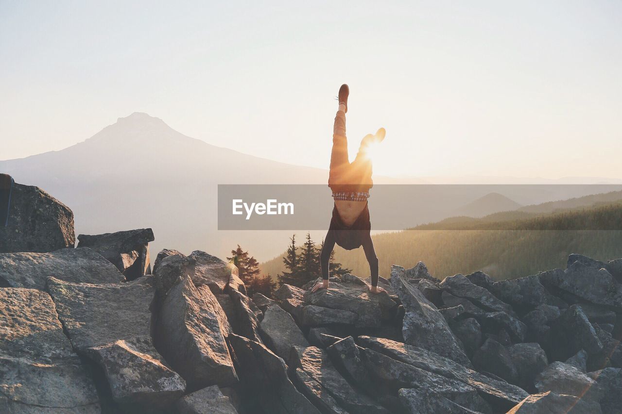 Man doing handstand on rocks against sky during sunrise