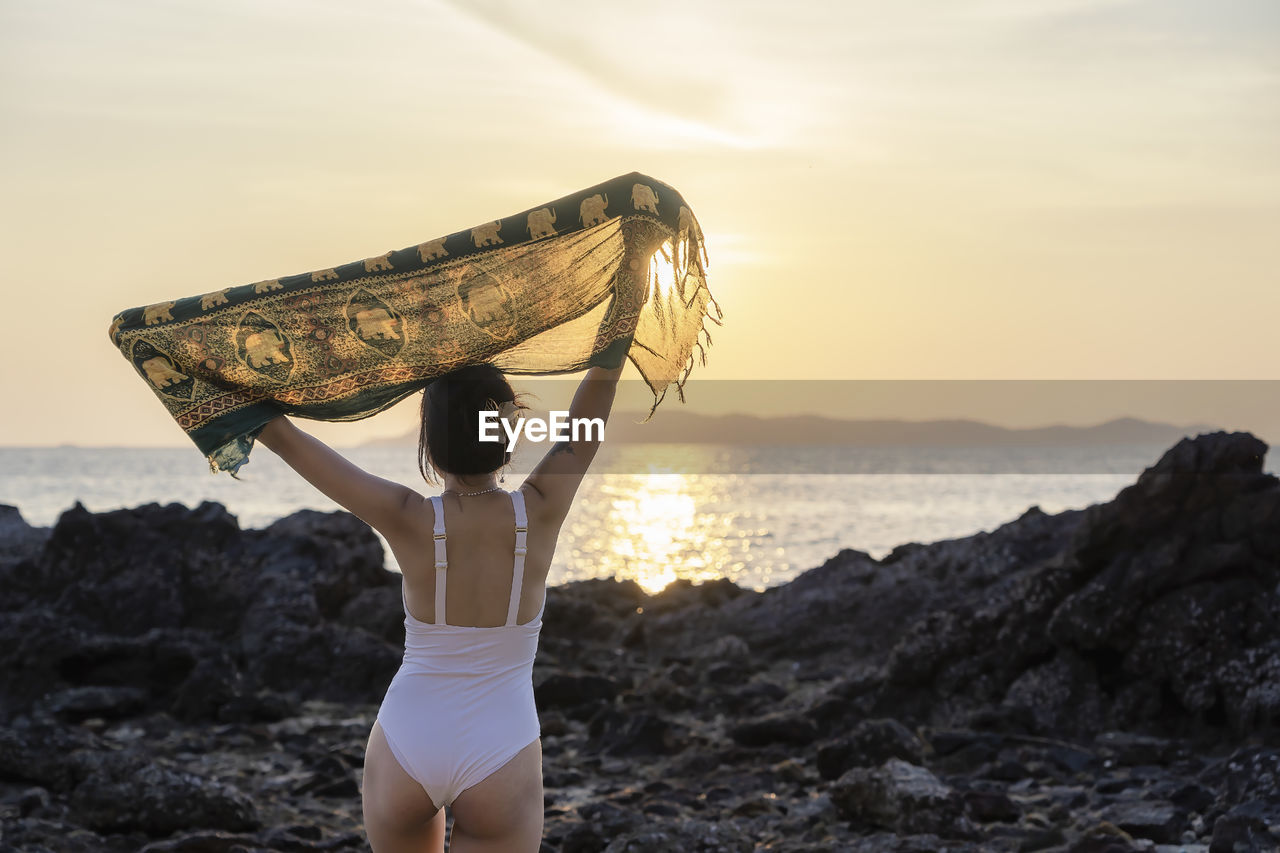 Woman tourist raising hands with sunset at beach on vacation trip. female relaxing with common cloth 