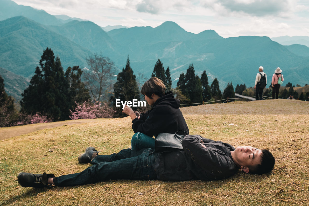 REAR VIEW OF PEOPLE RELAXING ON MOUNTAIN AGAINST SKY