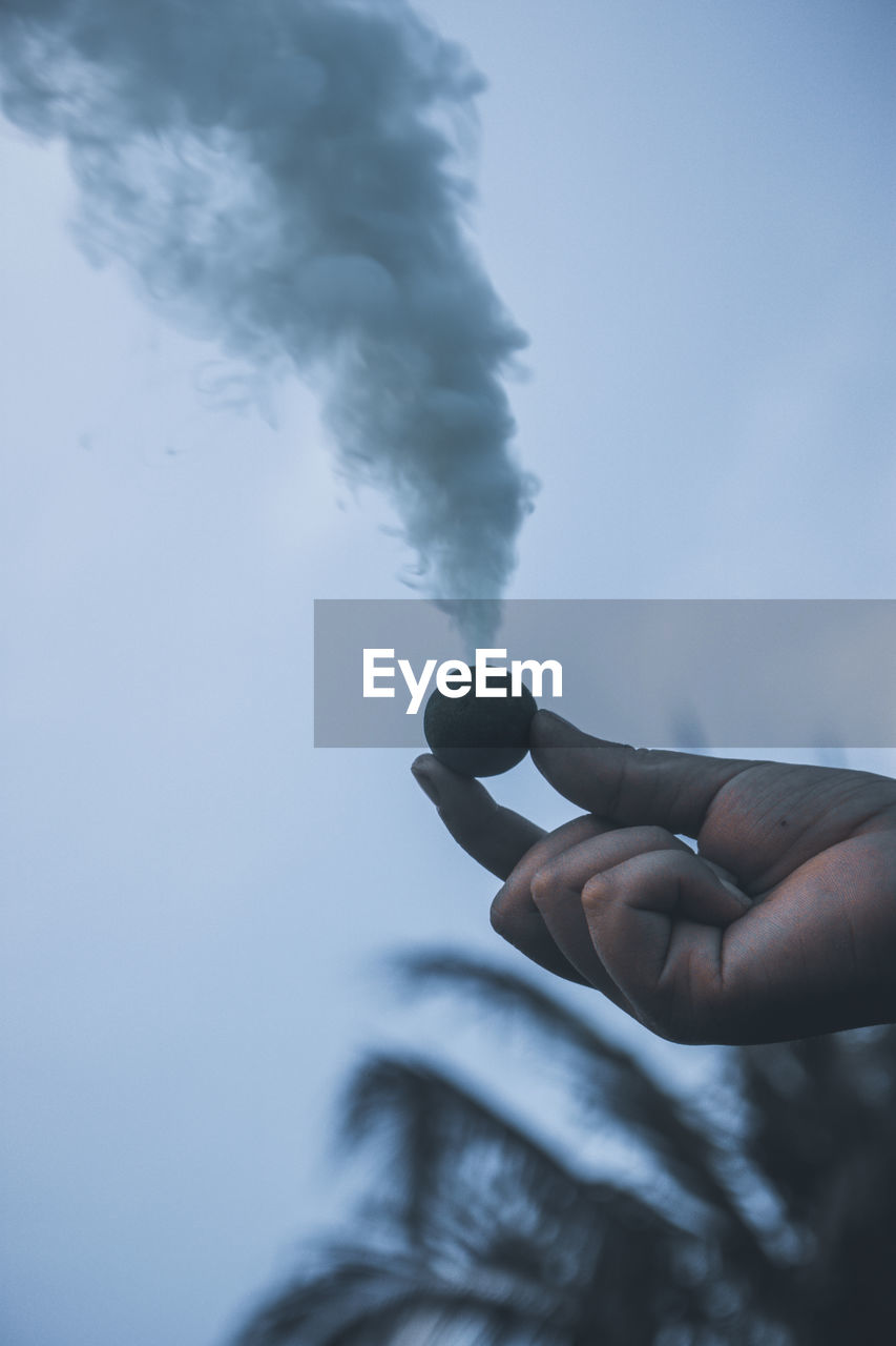 Cropped hand of person holding firework with smoke against sky