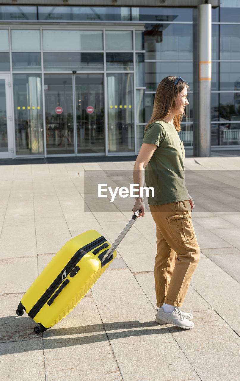 Young woman traveler carrying a yellow suitcase next to the entrance to the airport outside luggage